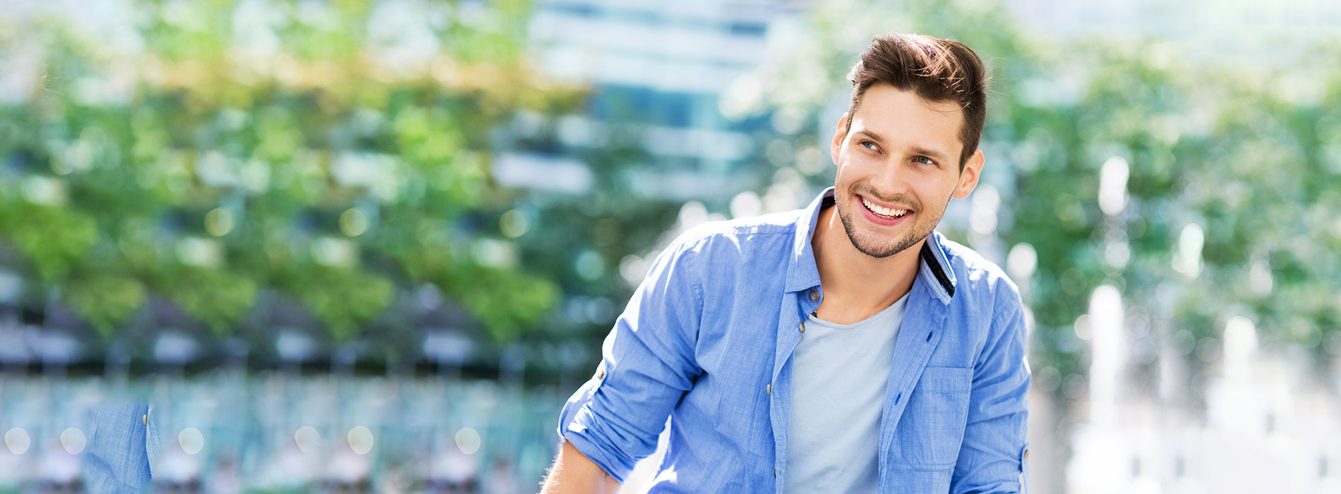 The image is a photograph of a young man with short hair, smiling at the camera. He is wearing a light blue shirt and is standing outdoors in front of a blurred background that suggests an urban environment, possibly near a body of water or a park, during daylight.